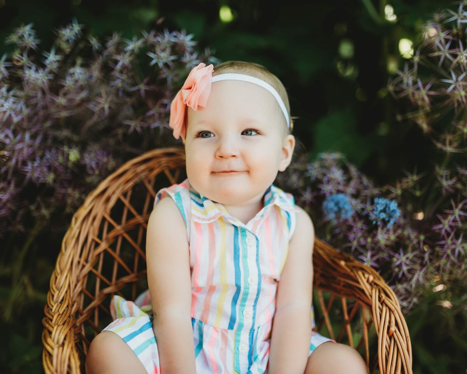 one year old girl twin sister in striped dress sitting on whicker chair flowers in the background