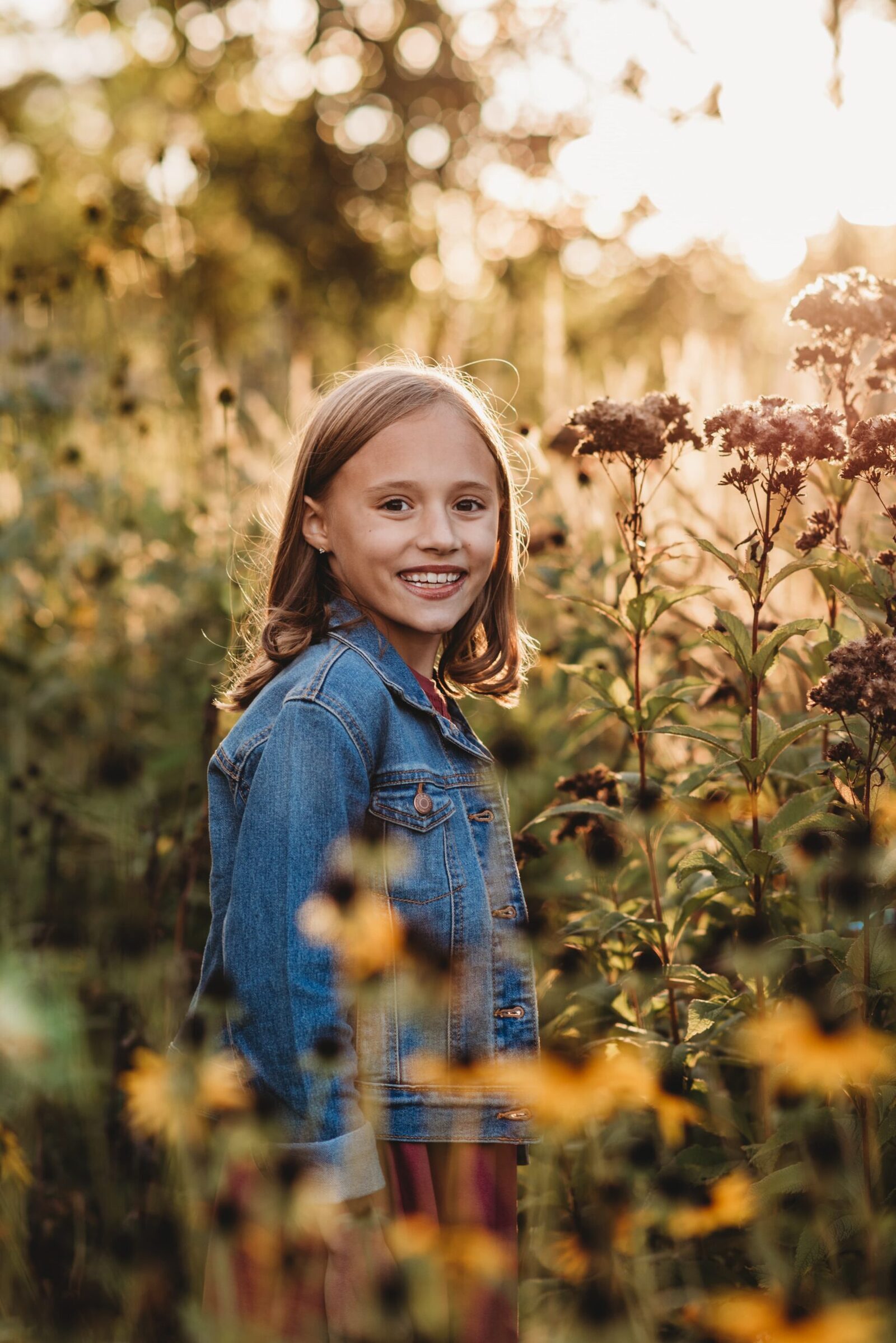 young girl in denim jacket standing in a flower garden with glowing sun in the background