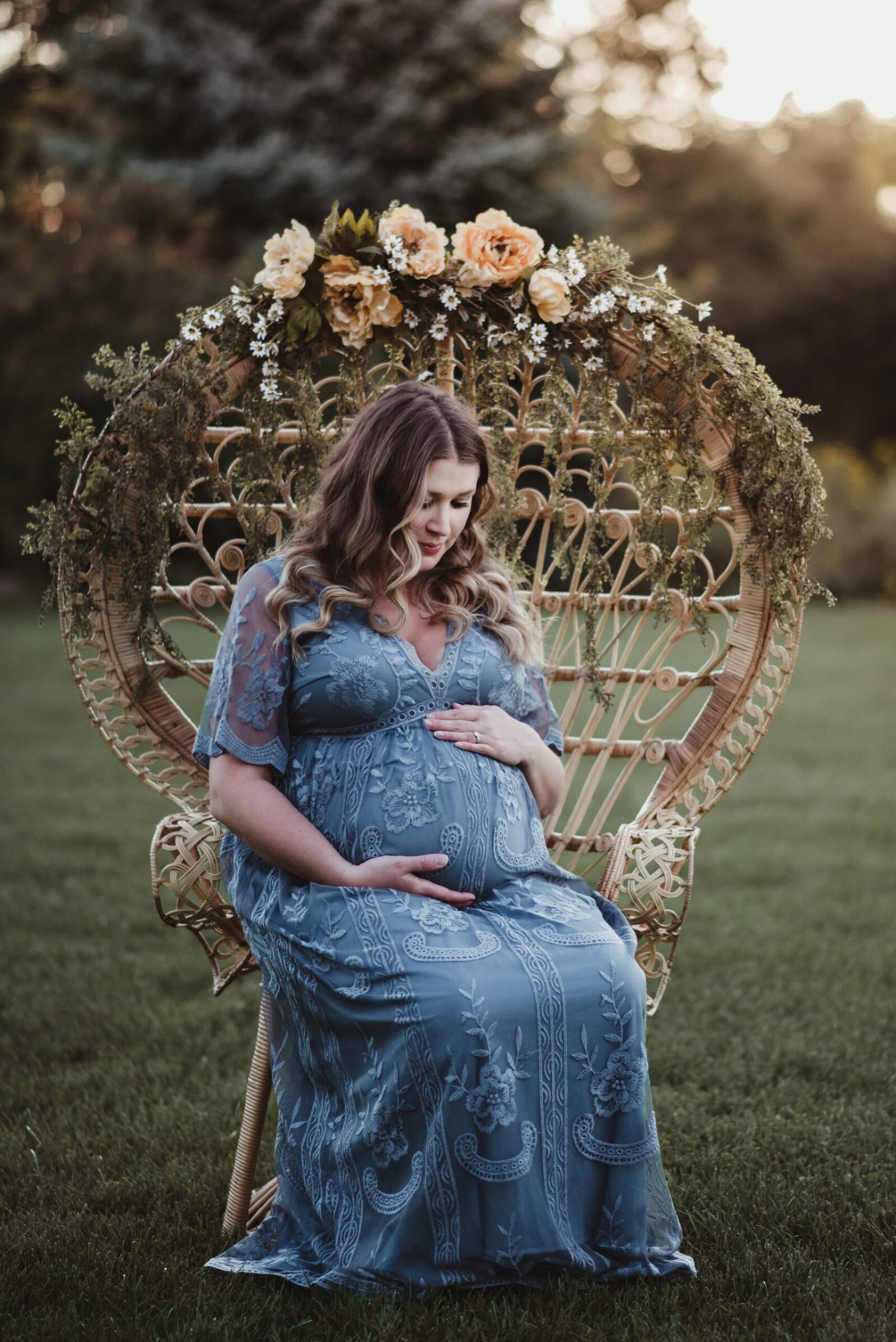 pregnant mother holding her belly in lacy blue dress sitting on peacock wicker chair decorated with flowers and greenery